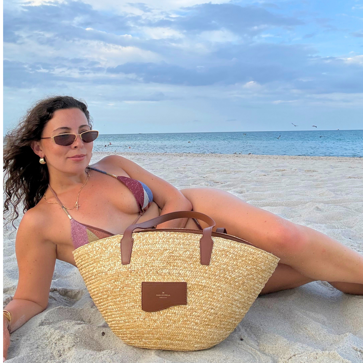 Lifestyle: Large Straw Marea Tote Bag in Smooth Brown leather (Chestnut brown), bag on sand in front of person. Size reference, beach scene.